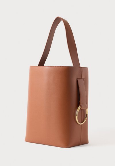 Hnedá vegánska kožená taška na nosenie, valcovitý tvar, ploché dno a zlatý kovový kruh ako ozdoba na boku. Pevný popruh.