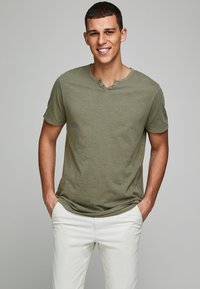 Oliven grøn bomulds t-shirt med rund halsudskæring, korte ærmer og tre knapdetaljer, parret med hvide bukser.