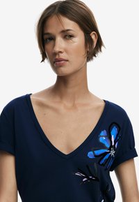 Navyblå V-ringad t-shirt i mjukt tyg, med livfulla blommönster i nyanser av blått och svart på vänster sida.