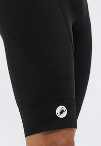 Κοντινό πλάνο μαύρων σορτς ποδηλασίας με ένα μικρό λευκό λογότυπο σκορπιού στην κάτω περιοχή του μηρού, πάνω σε απλό φόντο.