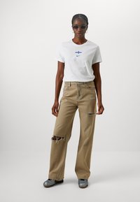 White T-shirt with blue graphic logo, paired with beige ripped wide-leg jeans. Model wearing small sunglasses and gray shoes.