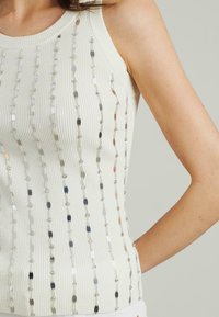 Top senza maniche bianco a coste con accenti di paillettes argento, oro e blu navy disposti verticalmente, con scollo rotondo e una texture morbida.