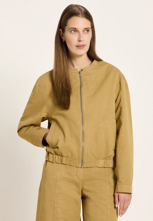 Frau mit langem, glattem Haar, die eine beigen Reißverschlussjacke und passende Hose trägt, leicht nach rechts schaut und eine Hand in der Hosentasche hat.