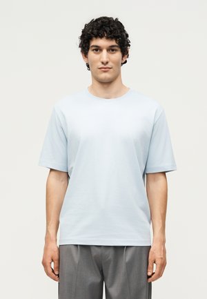 Tânăr cu păr creț, purtând o cămașă simplă albastru deschis cu mânecă scurtă și pantaloni gri, stând în fața unui fundal simplu.