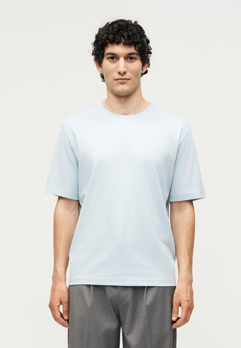 Νέος άνδρας με σγουρά μαλλιά που φοράει ένα απλό ανοιχτό μπλε κοντομάνικο πουκάμισο και γκρι παντελόνια, στέκεται μπροστά σε απλό φόντο.