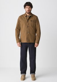 Brown cotton jacket with a button front, chest pocket, and side pockets, paired with dark denim jeans and beige suede shoes.