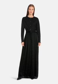 Schwarze Strickjacke mit rundem Ausschnitt und fünf Knöpfen, kombiniert mit einem schwarz gepleateten Rock mit vertikalen Streifen und einem taillierten Gürtel.