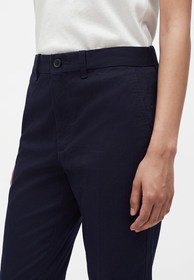 Personne portant un pantalon bleu marine foncé avec un bouton et des passants de ceinture, et une chemise blanche rentrée, la main droite reposant le long du corps.