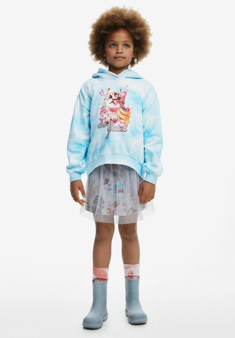 Lichtblauwe tie-dye hoodie met kattenafbeelding, gelaagd over een doorschijnende witte rok met kleurrijke patronen. Gedragen met grijze regenlaarzen en roze sokken.