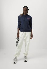 Μπλε-σκούρο φούτερ με ροζ λογότυπο, τσέπη καγκουρό, σε συνδυασμό με λευκή φόρμα και γκρι αθλητικά παπούτσια· κρατώντας σκοινάκι στο αριστερό χέρι.