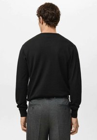 Man die met zijn rug naar de camera staat, draagt een zwart shirt met lange mouwen en grijze nette broek tegen een effen lichte achtergrond.