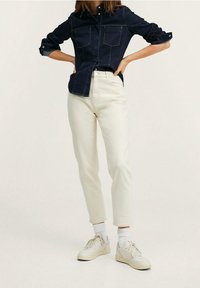 Camisa de mezclilla azul oscuro con botones y dos bolsillos en el pecho, combinada con jeans rectos beige claro y zapatillas blancas. Fondo liso.