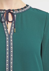 Tealfärgad blus med V-ringning och marinblå kant, dekorativa vävda detaljer och dragsko med guldspetsar.