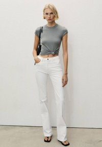 Tricou gri ajustat cu un detaliu legat la talie, combinat cu blugi albi evazați și sandale negre. Stil minimalist, modern.