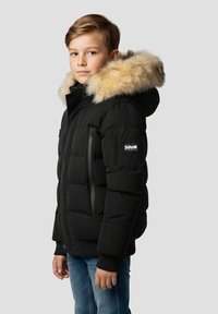 Sort polstret jakke med pelsforet hætte, lynlåslommer og et logopatch på ærmet. Båret af et barn, kombineret med blå jeans.