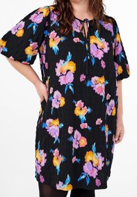 Femme portant une robe noire avec des motifs floraux violets, orange et bleus, et des manches bouffantes courtes, la main placée dans la poche latérale.