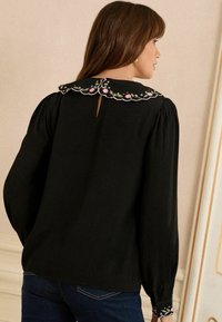 Blusa nera con colletto ricamato a scallop con motivi floreali in rosa e verde. Maniche lunghe con spalle arricciate e apertura a chiave sulla schiena.