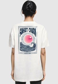 T-shirt bianco in cotone con una grafica sul retro che rappresenta un sole rosa con raggi sopra un'onda stilizzata, incorniciata dal testo "SUNSET CHARM" e "GOLDEN HOUR."