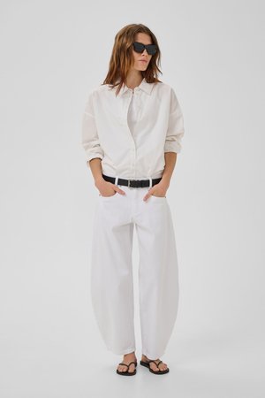 Žena v nadmernej bielej košeli na gombíky, bielych širokých nohaviciach s čiernym opaskom, čiernych sandáloch a tmavých slnečných okuliaroch stojaca s rukami v vreckách.
