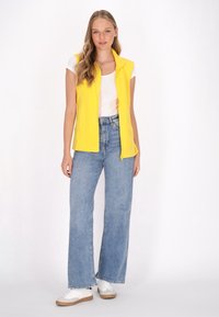 Gelbes ärmelloses Oberteil mit hohem Kragen, kombiniert mit hellblauen weit geschnittenen Jeans und weißen Sneakers. Schlichtes Design und lässige Passform.