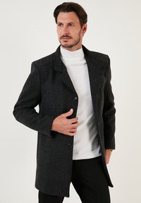 Abrigo negro de textura con un ligero brillo, con cuello de muesca, cierre de un solo botón y dos bolsillos frontales, llevado sobre un cuello de tortuga blanco.