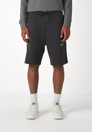 Shorts de sport noirs avec une taille à cordon de serrage, poches latérales et un logo vert. Associés à des baskets blanches et des chaussettes blanches mi-mollet.
