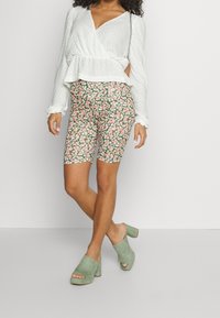 Nainen, joka on pukeutunut valkoiseen teksturoituun kääntöpaidasta, kukkakuosiset biker-shortsit ja salvia vihreät avokkaat, seisoo valkoista taustaa vasten.