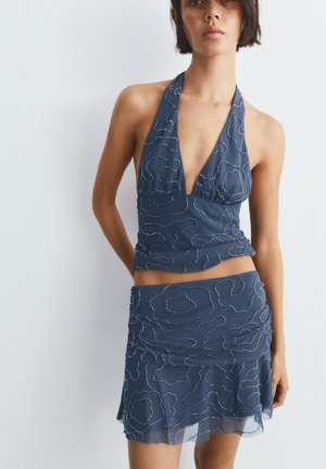 Žena nosiaca modrý halter top a zladenú sukňu s bielymi bodkovanými abstraktnými vzormi, stojaca na jednoduchom svetlom pozadí.