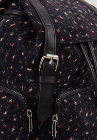 Gros plan d'un sac à dos en tissu noir avec de petits motifs floraux rouges et blancs, doté de fermetures éclair argentées et d'une sangle à boucle en cuir noir.