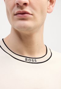 T-shirt en coton beige avec un col côtelé noir et un logo "BOSS" brodé en gris. Gros plan mettant en avant le design du col.