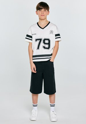 Chłopiec stoi z jedną ręką w kieszeni, ubrany w biało-czarną sportową koszulkę z numerem 79, czarne spodenki, skarpetki w paski oraz białe trampki.