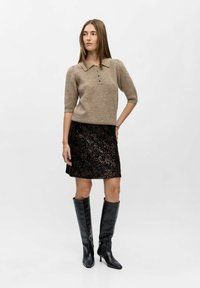 Beige wollen blouse met drie knopen, gecombineerd met een zwarte paillettenrok. Model draagt zwart leren laarzen tot de knie met hak. Eenvoudige achtergrond.