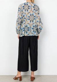 Blusa beige fluida con motivo floreale blu e nero, maniche lunghe. Abbinata a pantaloni neri a pieghe con gamba larga. Sandali con tacco a blocco marroni.