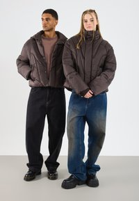 Două persoane îmbrăcate în jachete pufoase negre, blugi largi și pantofi cu platformă stau în fața unui fundal deschis.