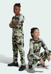 Tute in tessuto mimetico nei toni del verde e del marrone, con dettagli a striscia bianca. Un modello sta in piedi con le braccia incrociate, mentre l'altro è accovacciato.