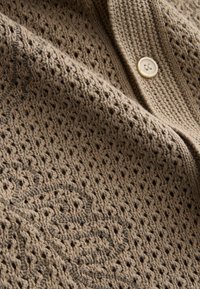 Beige strikket cardigan med et struktureret, åbent vævemønster. Har en rund, cremefarvet knap og diskret broderet detalje.