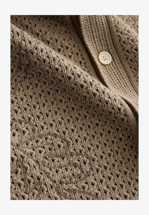 Beige gestrickte Strickjacke mit einem strukturierten, offenen Webmuster. Sie verfügt über einen runden, cremefarbenen Knopf und eine dezente gestickte Verzierung.
