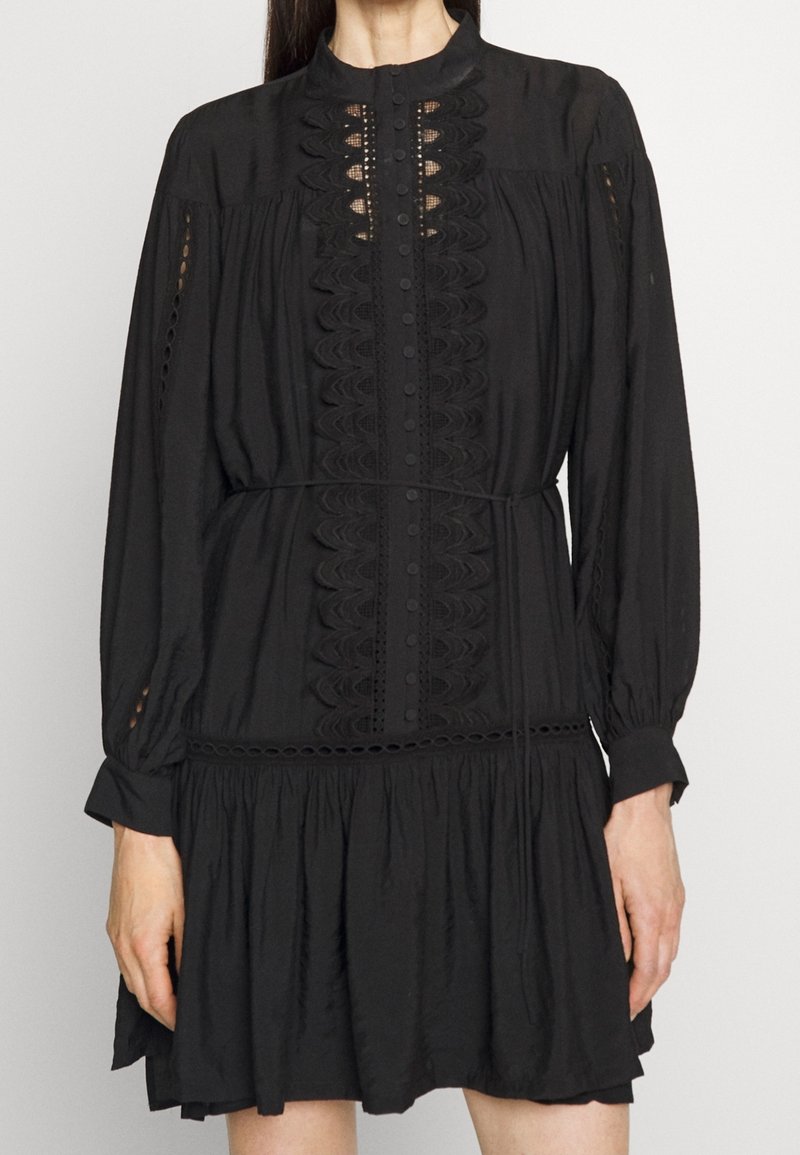 Femme portant une robe noire à manches longues avec des détails en dentelle, des boutons sur le devant, une taille cintrée et une jupe plissée au-dessus du genou.
