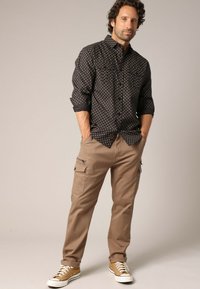 Zwarte overhemdd met gouden stippen, gecombineerd met khaki cargobroeken met zakken. Bruine schoenen met witte zolen maken de outfit compleet.