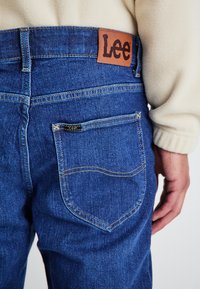 Vaqueros de mezclilla azul oscuro con un parche de cuero marrón con el logo de Lee, realzados con costuras amarillas y un pequeño detalle en el bolsillo trasero.