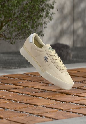 Beige leather sneaker with white accents, lace-up design, textured upper, and logo detail on the side. Lightweight sole for traction.