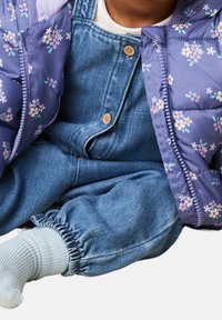 Dziecko w niebieskich dżinsowych ogrodniczkach, jasnoniebieskich skarpetkach i fioletowej kurtce w kwiatowy wzór, siedzące z crossed legs.