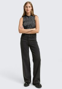 Młoda kobieta stoi ze skrzyżowanymi ramionami, ubrana w bezrękawnik z ciemnym motywem w panterkę, czarne szerokie dżinsy z wysokim stanem oraz czarne botki.