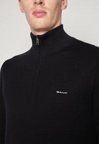 Zwarte gebreide trui met een hoge kraag, voorzien van een korte rits en een klein wit logo op de borst. Geweven stof met een aansluitende stijl.