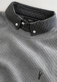 Grå stickad tröja med ribbad struktur, svart rutmönstrad knappad krage och en liten broderad hjortlogotyp.