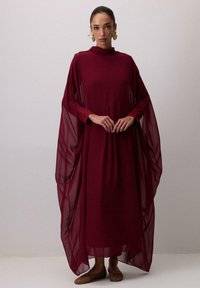 Burgundska duga haljina s prozirnim plaštastim rukavima i visokim ovratnikom, izrađena od lagane tkanine. Naglašena plisiranim dizajnom i ravnim cipelama.