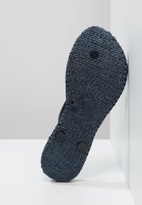Suola di scarpa nera con texture e design intrecciato, caratterizzata da due bottoni circolari neri. La suola presenta una leggera curva e un bordo piatto.