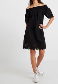 Černé šaty s odhalenými rameny a krátkými rukávy, mají detailování z krajkového vyšívání na lemu, vyrobené z lehké látky. Nošeny s bílými teniskami.