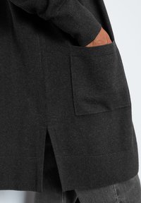 Mörkgrå cardigan i ett mjukt, texturerat material med en sidoklyvning och en framtida ficka på vänster sida.