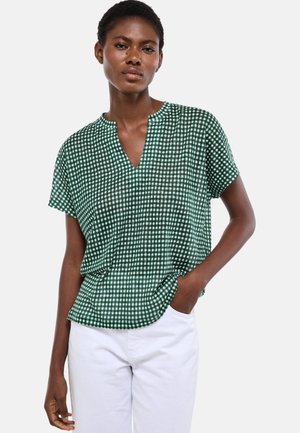 Femme portant un chemisier à manches courtes à carreaux verts et blancs avec un col en V, associé à un pantalon blanc taille haute, posant avec une main dans la poche.